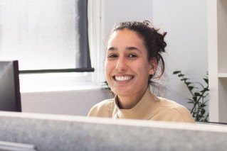Close-up of a young employee sitting behind a hotel reception, smiling directly into the camera.