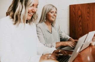 Zwei lachende Mitarbeitende arbeiten nebeneinander an ihren modernen Laptops.